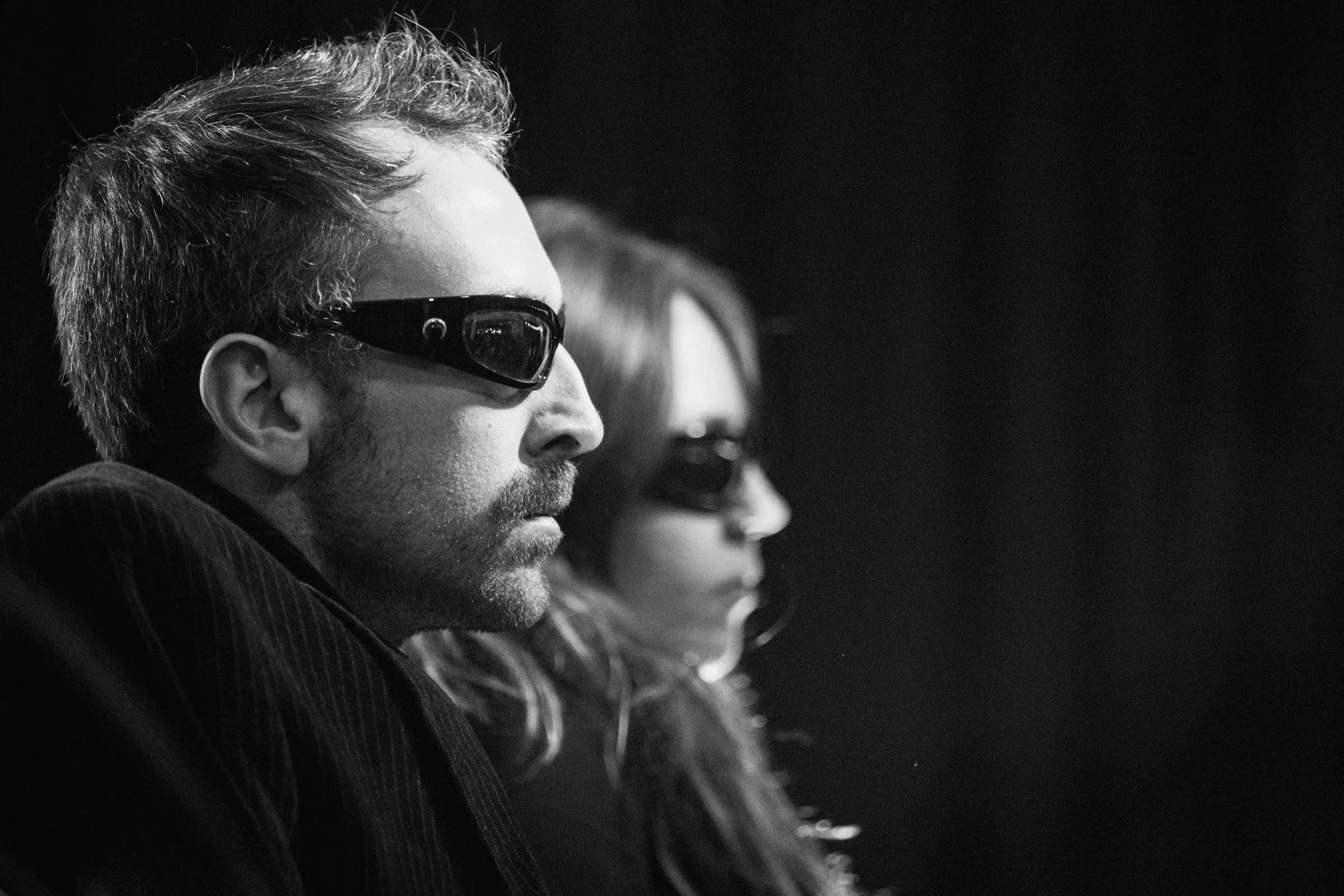 Black and white close-up. One member in wraparound sunglasses in profile in the foreground, the other out of focus behind him, against a dark stage.