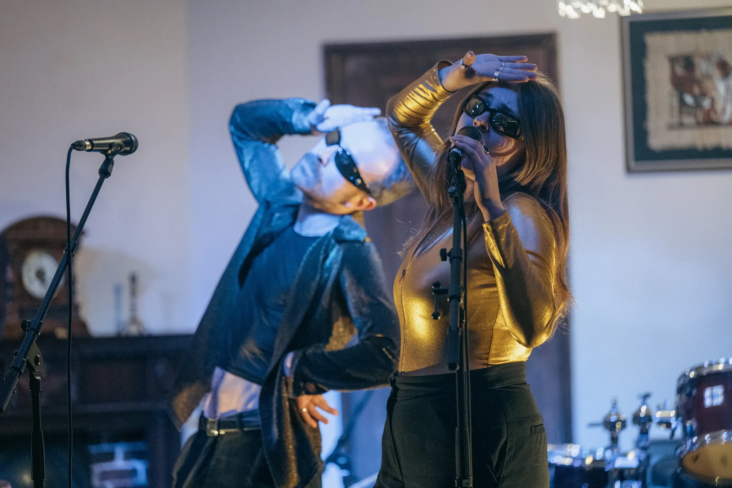 Two singers at microphones in a room with a mantel clock and a framed print behind them, one in a gold top shielding her eyes, the other leaning back with a hand raised.