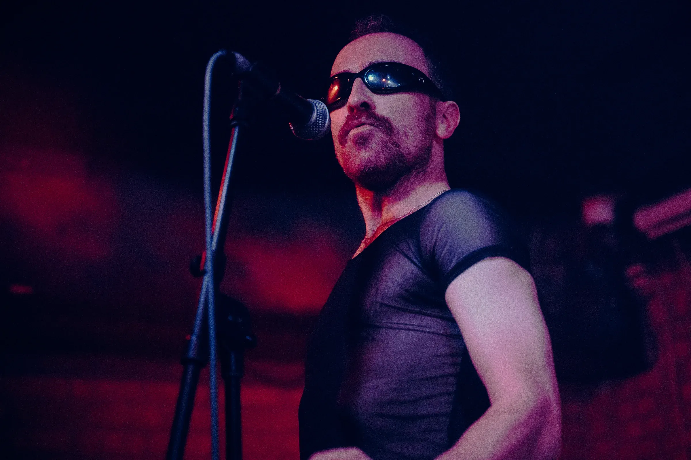 A singer in wraparound sunglasses and a black mesh top sings into a microphone, lit deep red from the side.