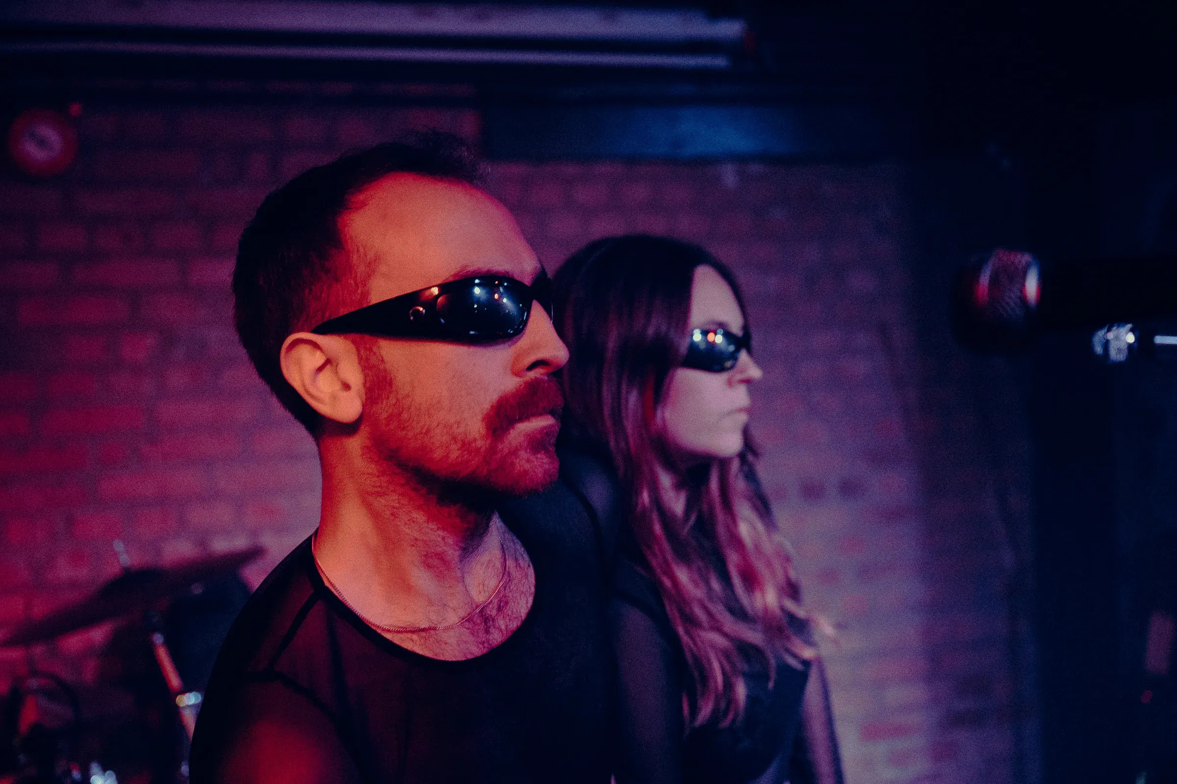 Both members in profile against an exposed brick wall under pink light, the edge of a drum kit visible at the left.