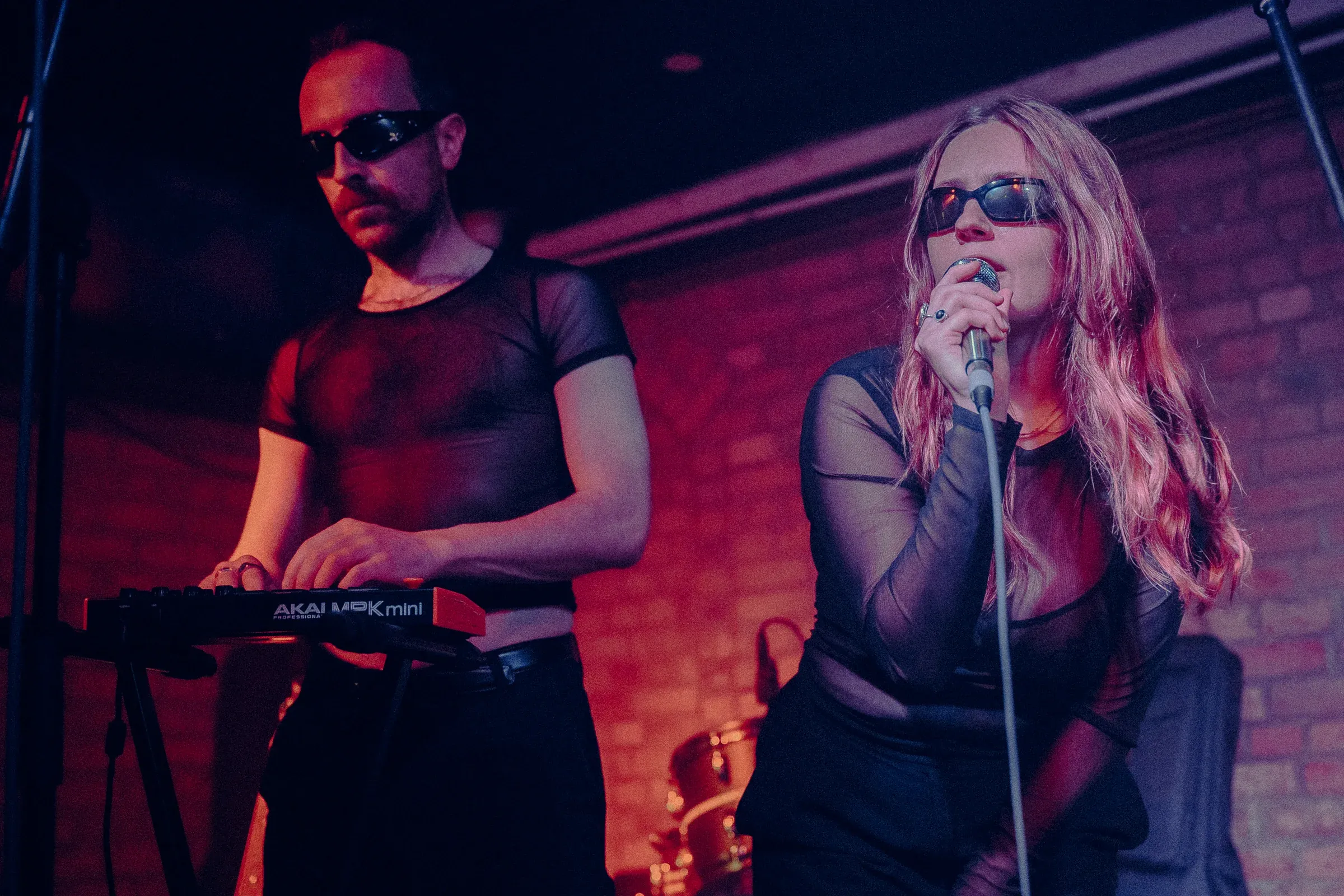 The pair on stage under red light, one playing a small Akai keyboard, the other singing into a microphone, both in dark glasses.