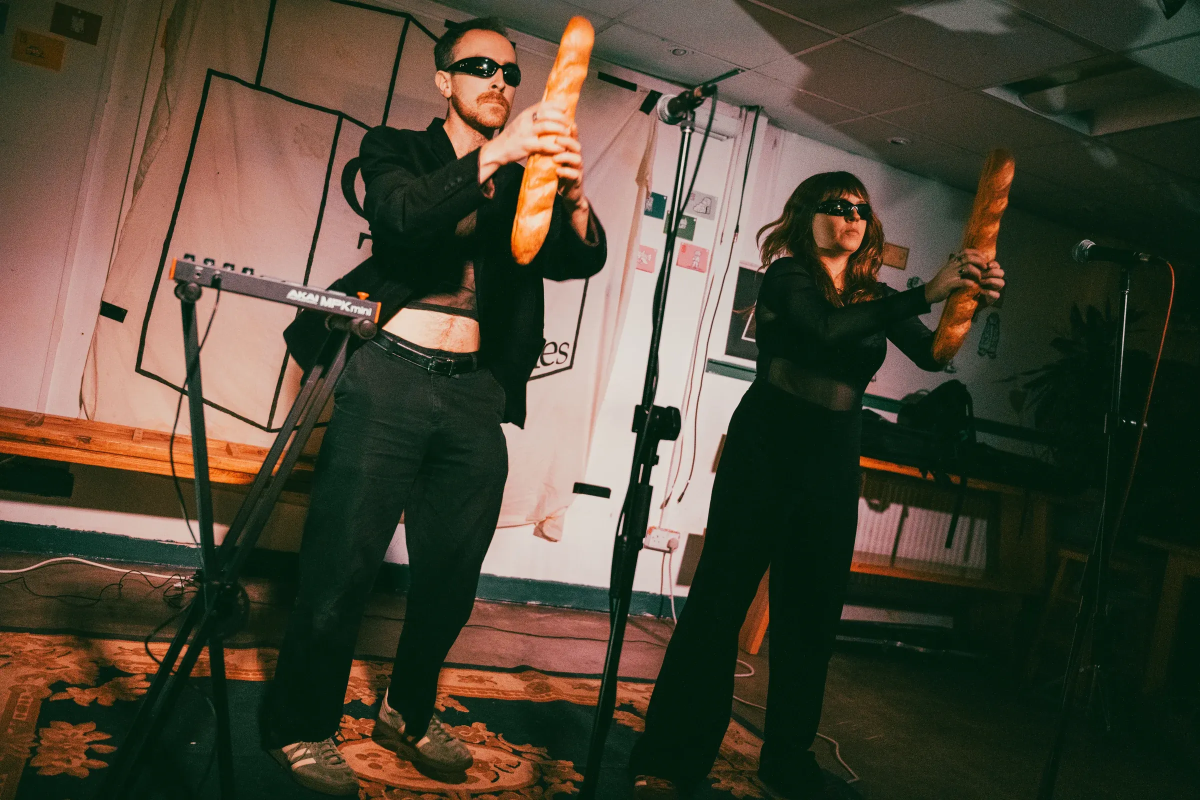 Both members of the band hold baguettes above their heads like weapons on a low stage in a function room, a patterned carpet under their feet and a keyboard on a stand beside them.