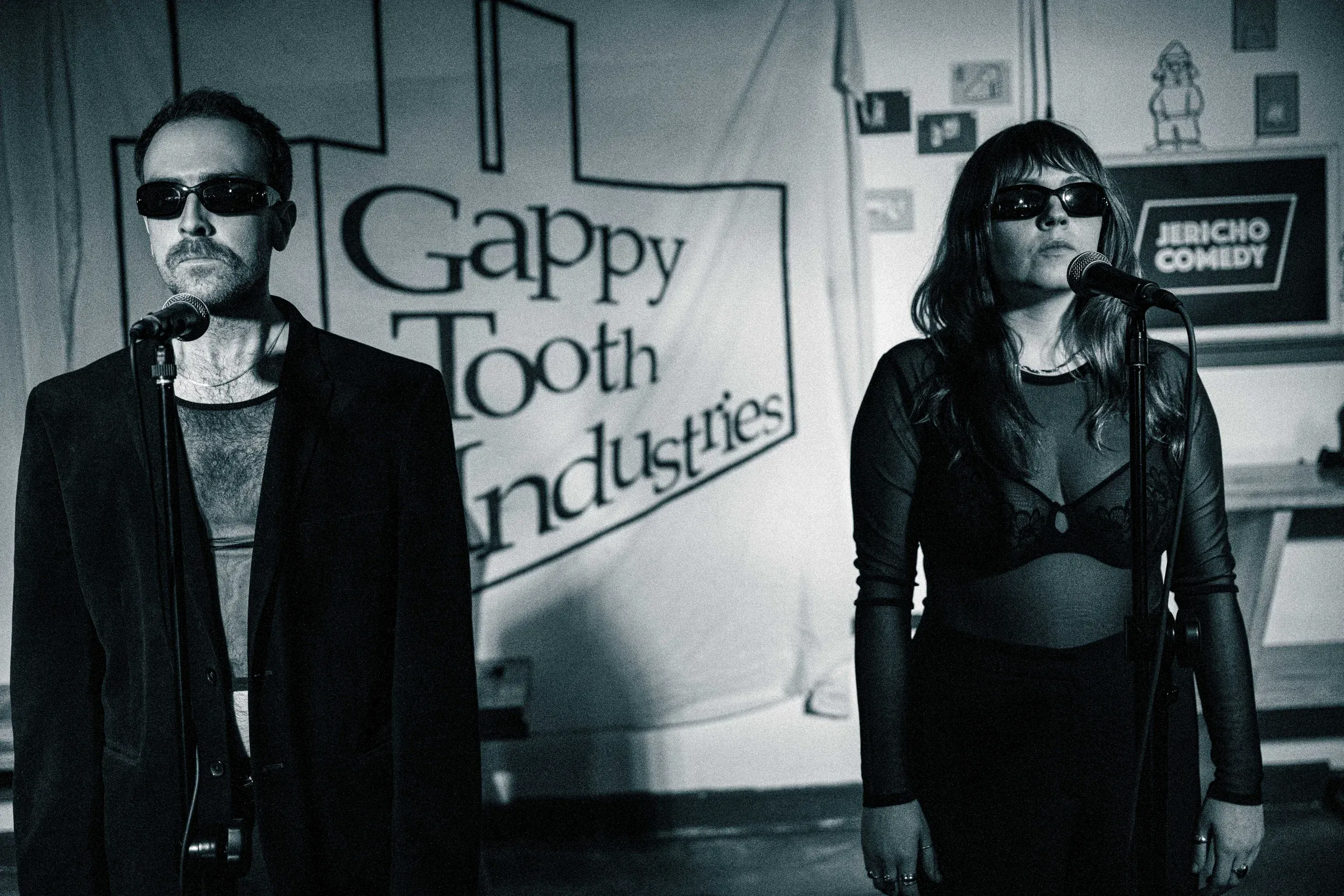 Black and white. The pair stand at separate microphones in front of a Gappy Tooth Industries banner, both wearing dark glasses.