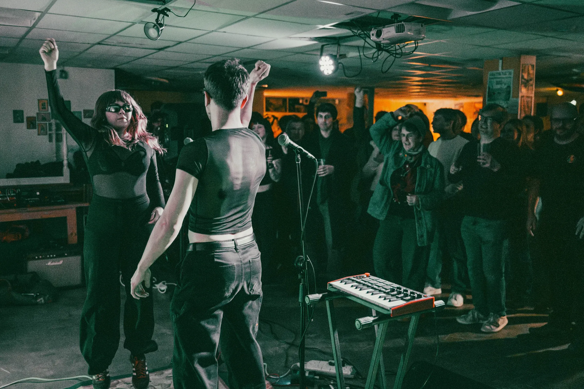 Seen from behind the stage, the pair face a crowd in a low-ceilinged room with their arms raised, a keyboard and microphone stand in the foreground.