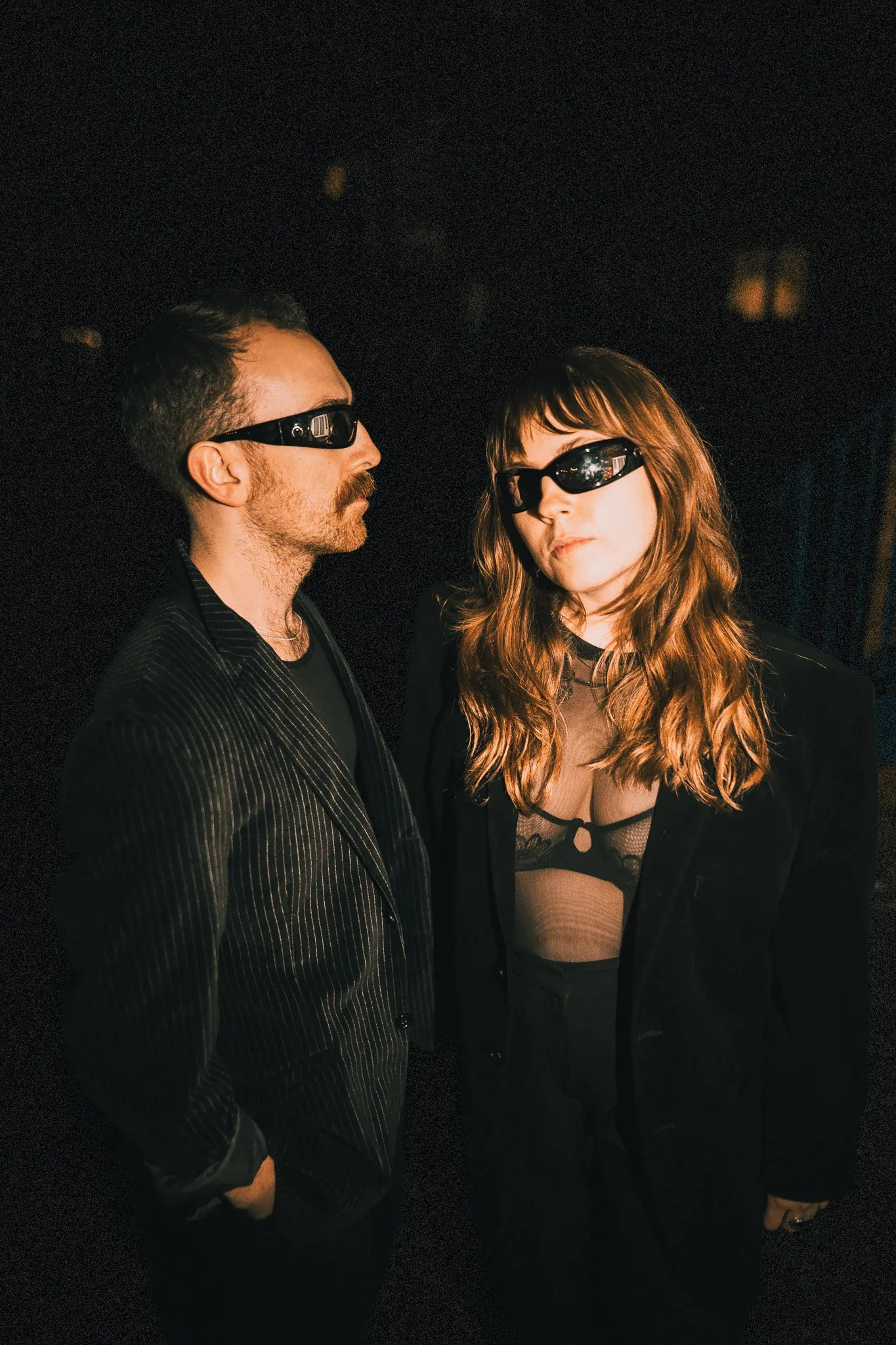 The two members of No Worries If Not outdoors at night in dark glasses and black tailoring, one looking towards the other.